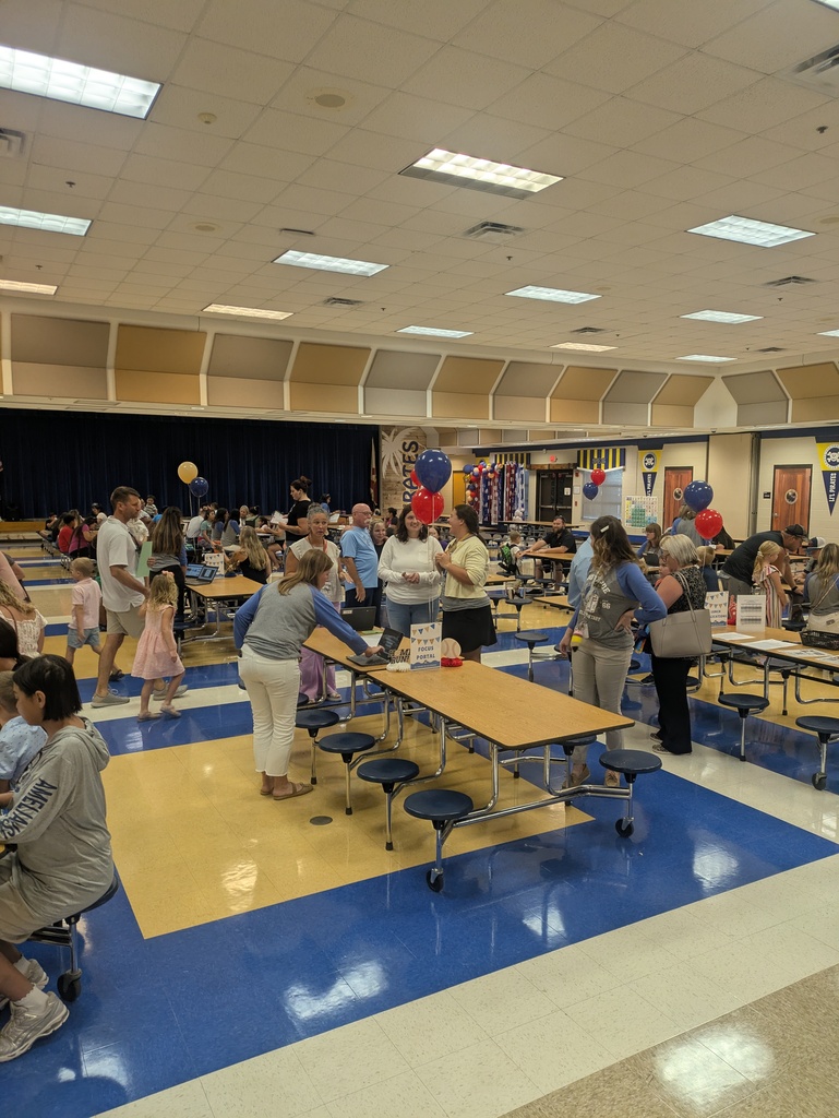 Students, families, and educators gather in a school cafeteria for a family event. Tables are decorated with blue, gold, and red balloons, and adults and children participate in activities throughout the space.