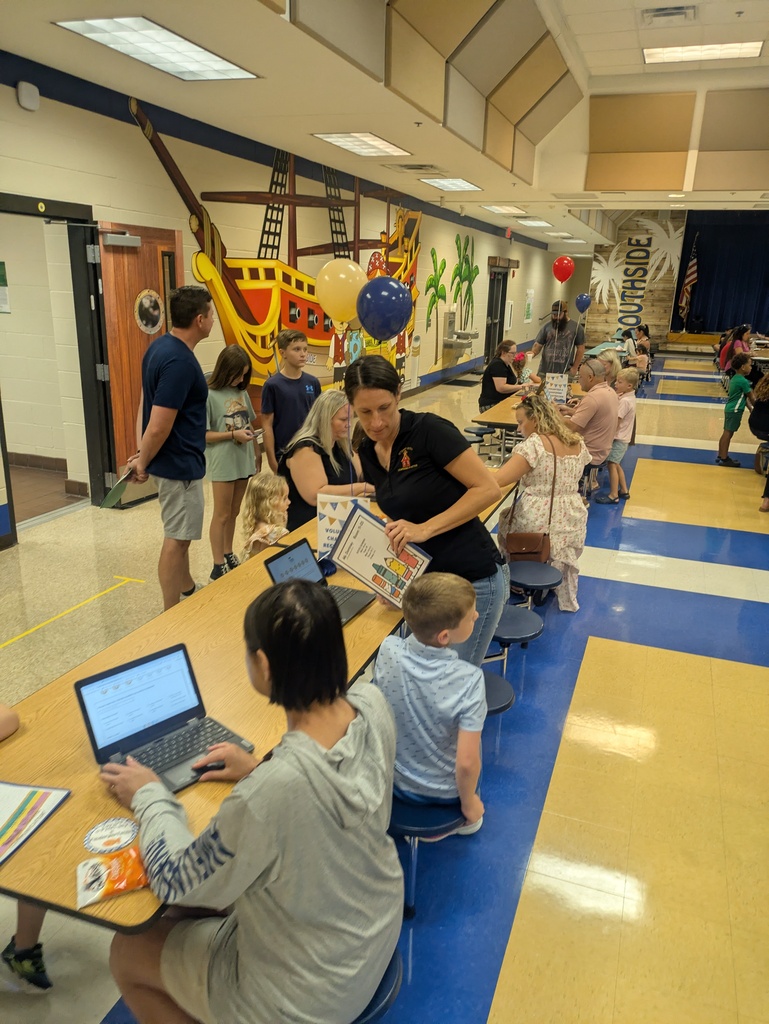 Students, families, and educators gather in a school cafeteria during a family event. Children hold activity materials while adults assist them at tables decorated with blue and gold balloons. A pirate-themed mural and school decorations are visible in the background.