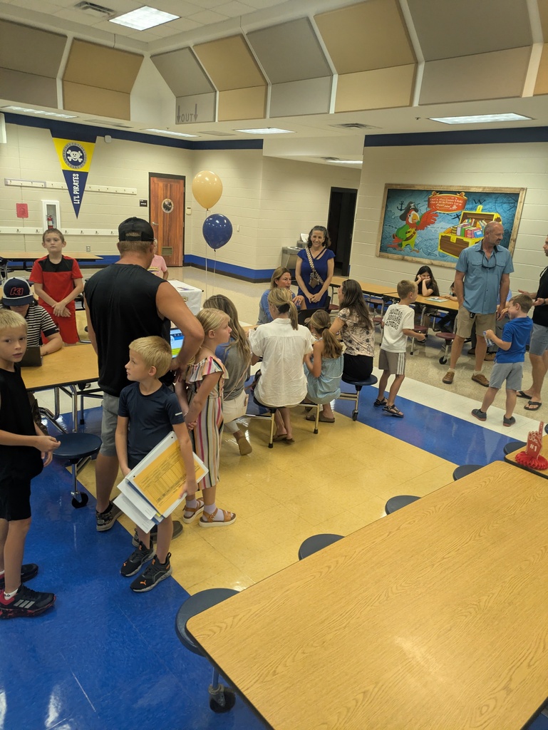 Families and educators sit together at cafeteria tables while participating in a school event. A pirate-themed mural, colorful decorations, laptops, and activity materials are visible throughout the space.