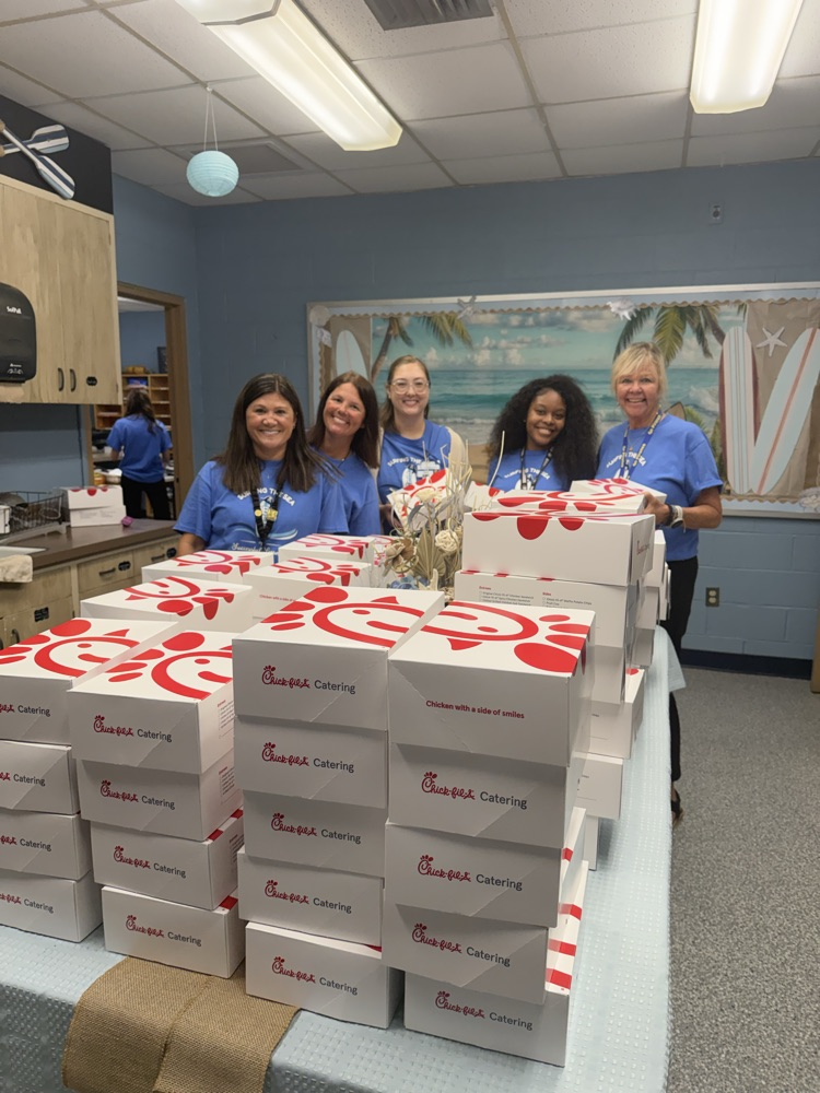 ELH staff smile behind a table full of chick-fil-a boxed lunches. The staff are wearing blue “Surf the Sea of Successful Learning"
