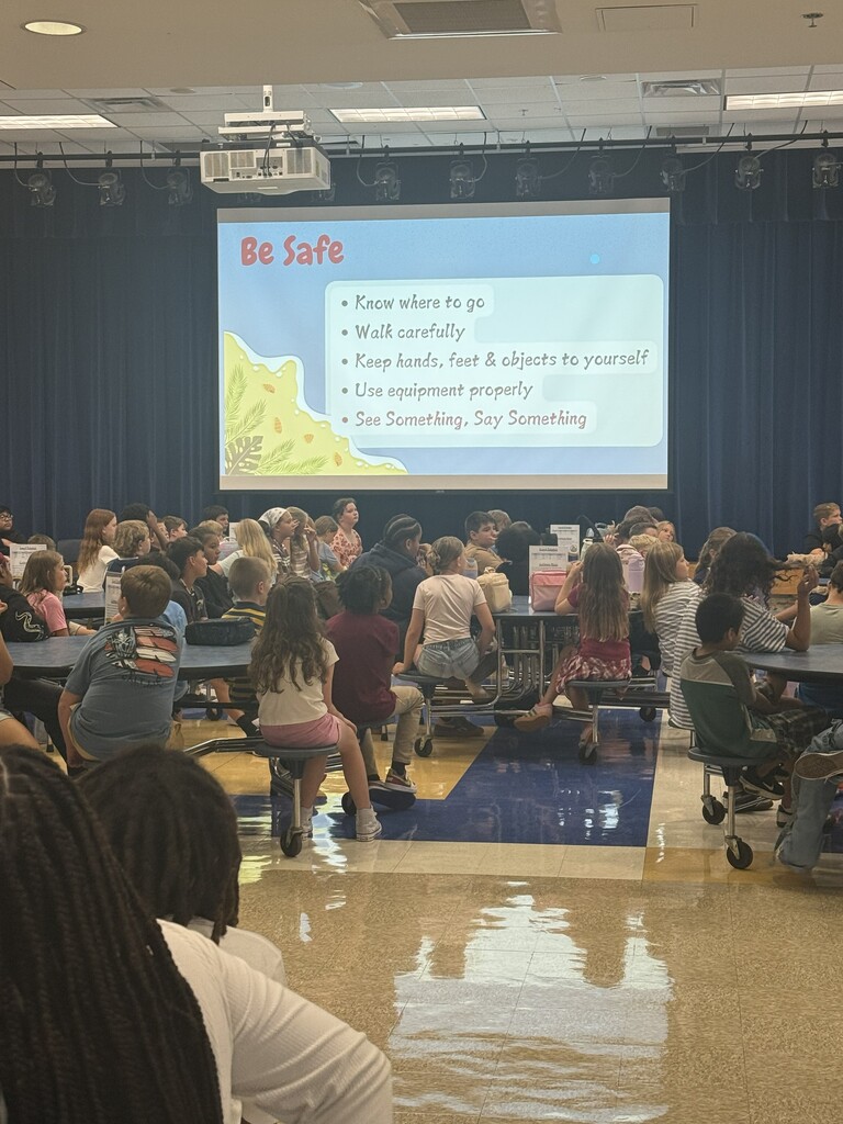 Students and teachers at a back to school pep rally in the cafeteria. The slide showing says "be safe".