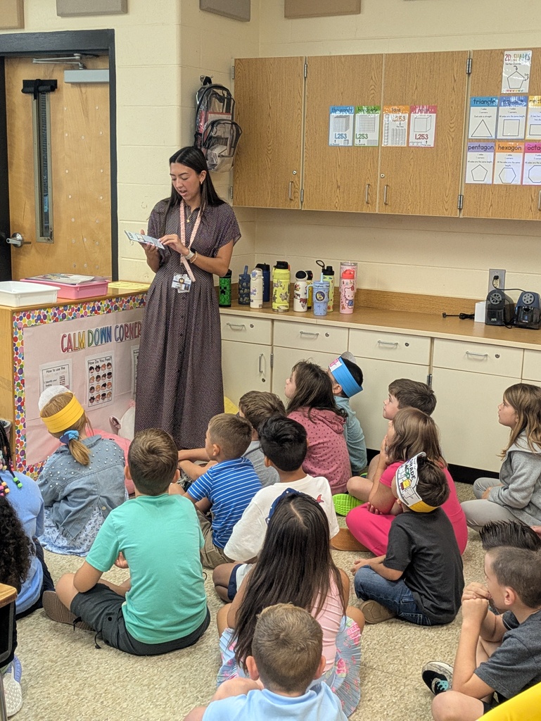 A teacher stands in front of a group of students sitting on the floor as she reads from a paper in her hand.