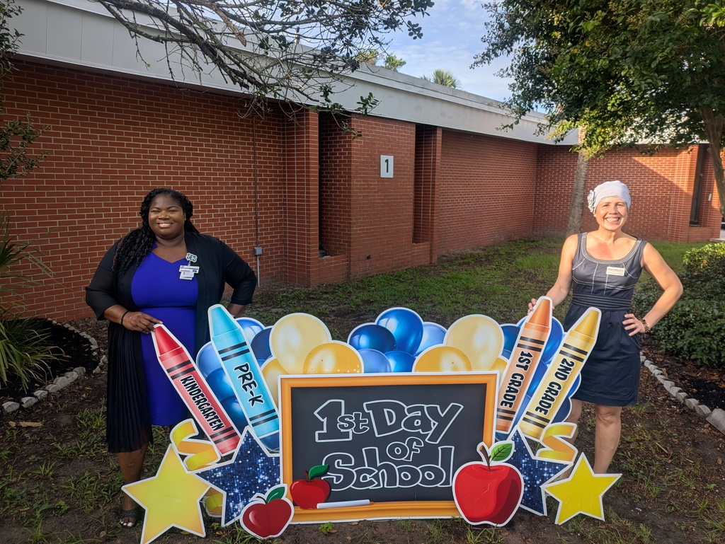 Two women stand to the left and right of a 1st day of school yard card sign that has gold and blue balloons with crayons that have the grade leveks, Pre-K, Kindergarten, 1st, and 2nd grade on them.