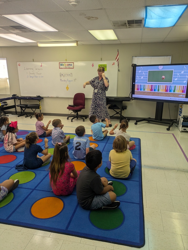 A teacher stands in front of a group of students sitting on carpet on the floor facing the teacher.