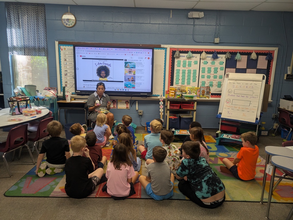 A group of students are sitting at the carpet of their classroom with their backs to the camera facing their teacher who is sitting holding an empty jar.