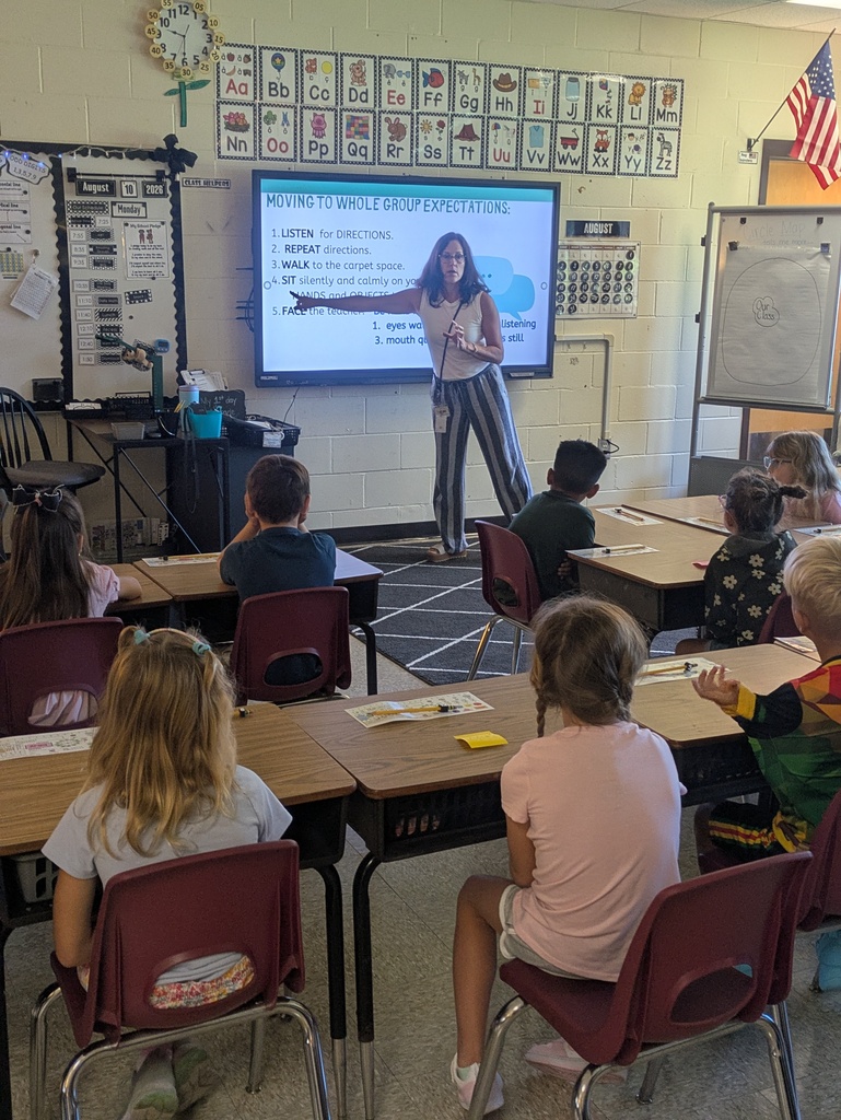 A teacher stands in front of students sitting at their desk pointing to her board that has whole group expectations written on them. 