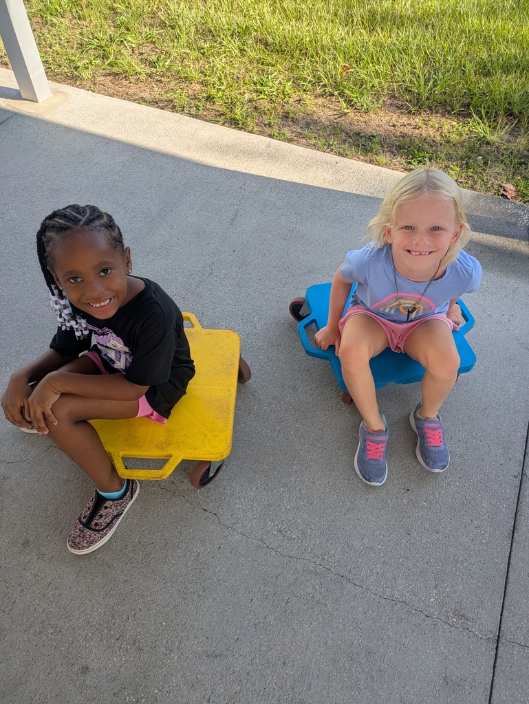 Two students sit on scooter boards one yellow and one blue  facing and smiling upward towards the camera.