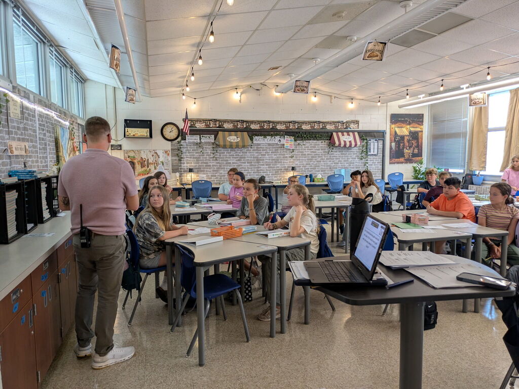 Students sitting at desks in a classroom looking at their teacher as he demonstrates a task for them.
