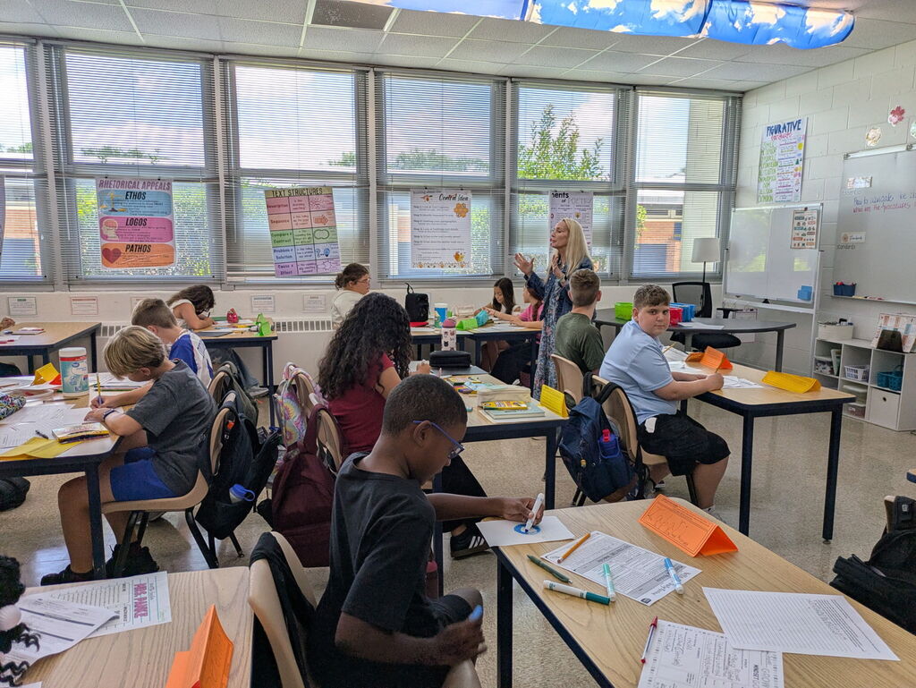 Students sitting at tables in a classroom while the teacher gestures with her hands.