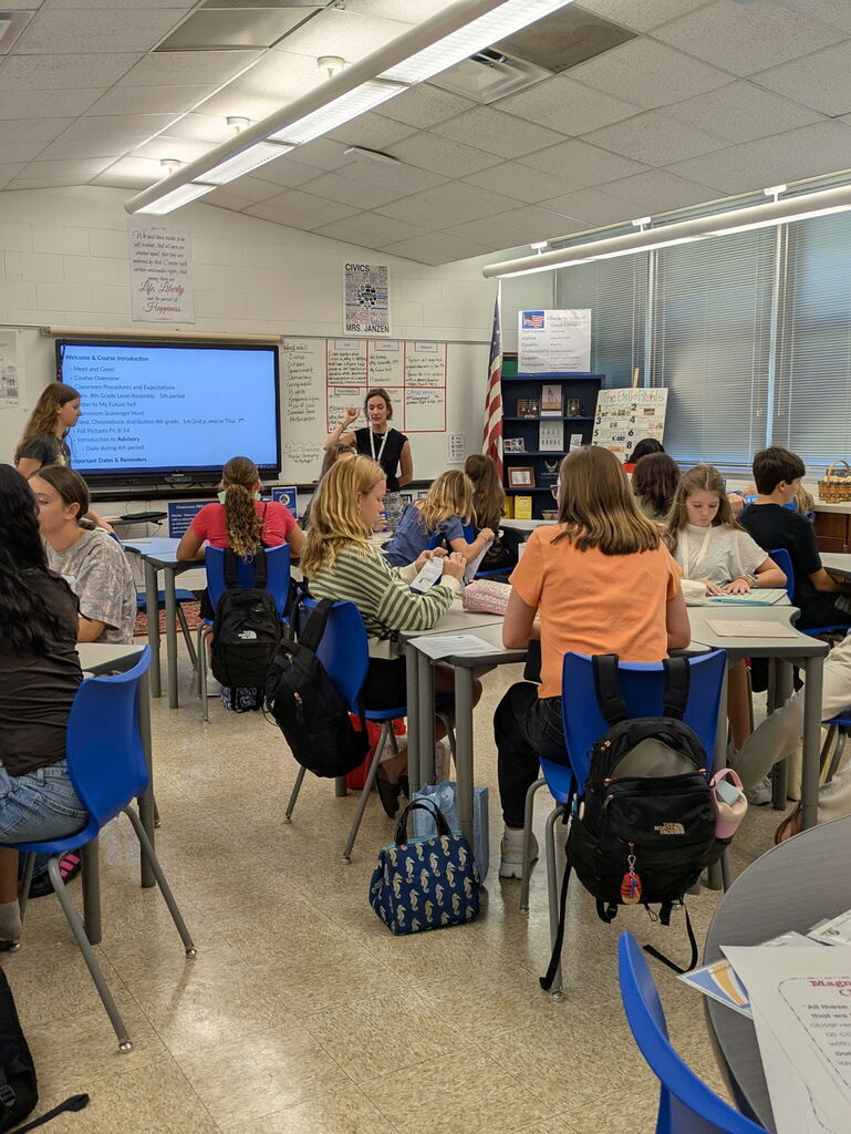 Picture of a classroom with a teacher pointing to a smart screen and students sitting at desks turned toward the teacher. 