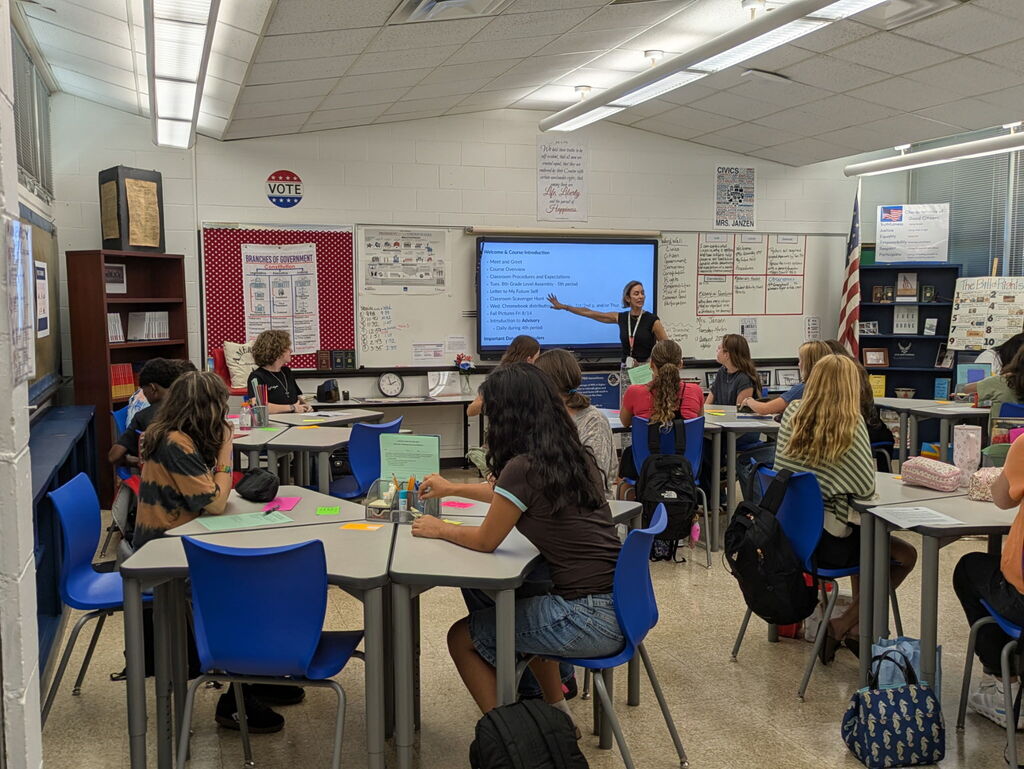 Picture of a classroom with a teacher pointing to a smart screen and students sitting at desks turned toward the teacher. 