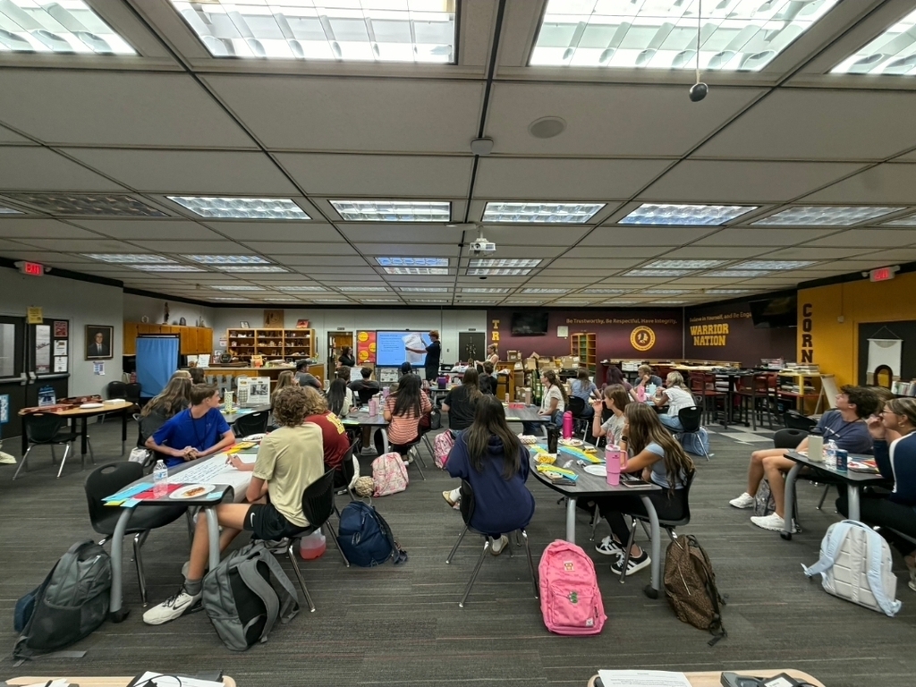 A series of photos shows West Nassau High School students actively participating in a collaborative classroom lesson. Students work in small groups around tables, discussing ideas, writing responses on large chart paper, and sharing their thinking with classmates. Several students stand at the front of the room to present their group’s work while their peers listen. Other photos capture students moving around the classroom and library, collaborating with different classmates during the activity. The learning spaces feature West Nassau’s garnet and gold colors, Warrior Nation signage, TRIBE expectations, classroom displays, bookshelves, and student materials. The photos highlight student engagement, teamwork, discussion, movement, and collaborative learning in action.