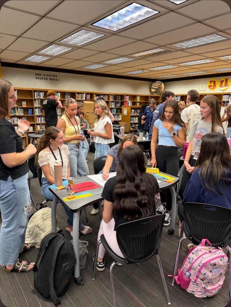 A series of photos shows West Nassau High School students actively participating in a collaborative classroom lesson. Students work in small groups around tables, discussing ideas, writing responses on large chart paper, and sharing their thinking with classmates. Several students stand at the front of the room to present their group’s work while their peers listen. Other photos capture students moving around the classroom and library, collaborating with different classmates during the activity. The learning spaces feature West Nassau’s garnet and gold colors, Warrior Nation signage, TRIBE expectations, classroom displays, bookshelves, and student materials. The photos highlight student engagement, teamwork, discussion, movement, and collaborative learning in action.