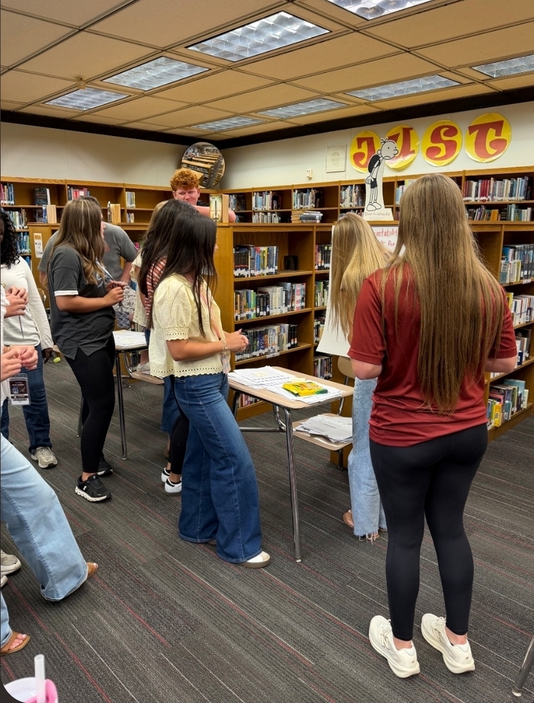 A series of photos shows West Nassau High School students actively participating in a collaborative classroom lesson. Students work in small groups around tables, discussing ideas, writing responses on large chart paper, and sharing their thinking with classmates. Several students stand at the front of the room to present their group’s work while their peers listen. Other photos capture students moving around the classroom and library, collaborating with different classmates during the activity. The learning spaces feature West Nassau’s garnet and gold colors, Warrior Nation signage, TRIBE expectations, classroom displays, bookshelves, and student materials. The photos highlight student engagement, teamwork, discussion, movement, and collaborative learning in action.