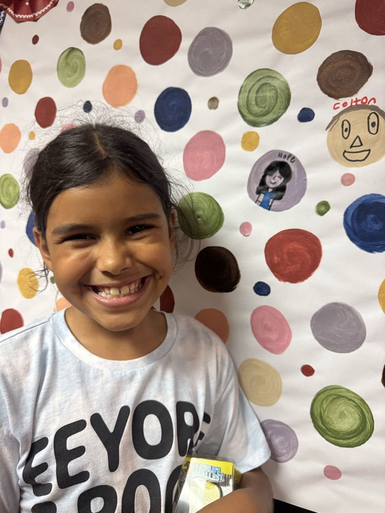 A smiling student proudly stands beside her artwork displayed on a colorful school bulletin board. Her creative portrait is featured among painted circles, celebrating her artistic expression and contribution to the display.