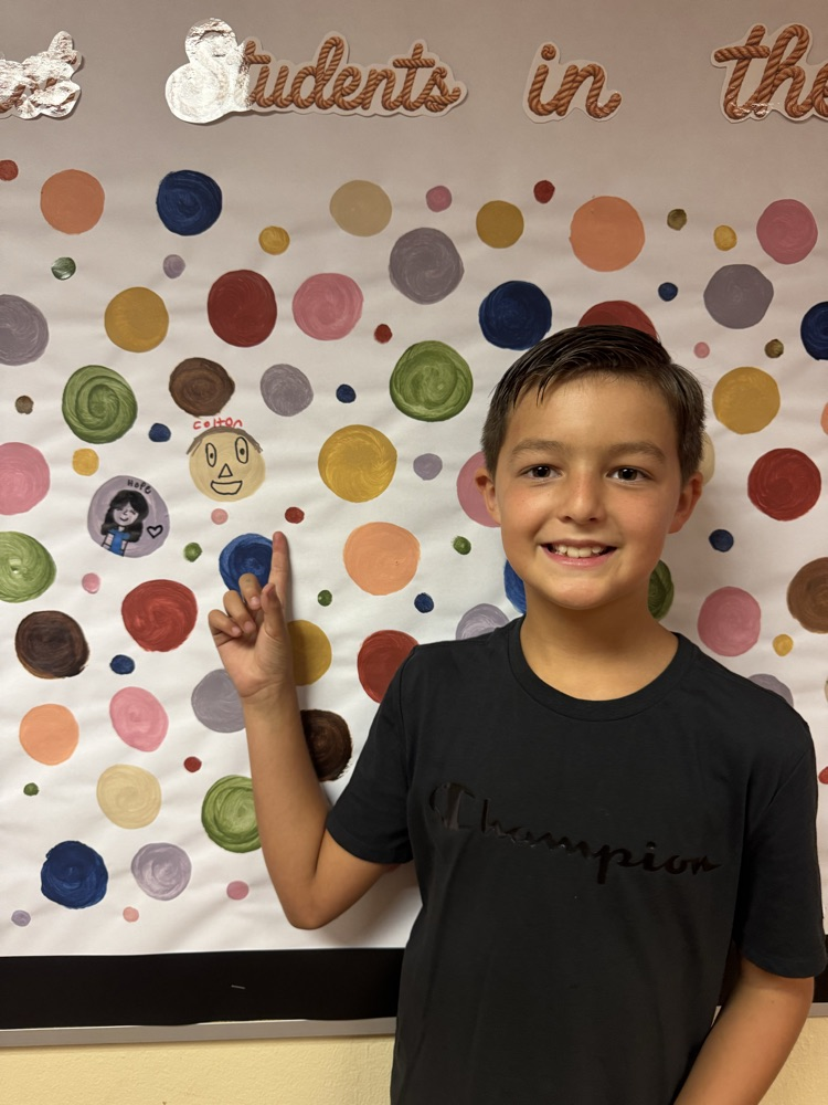 A smiling student proudly points to his artwork displayed on a colorful school bulletin board. His creative portrait is showcased among painted circles, highlighting student creativity and artistic expression.