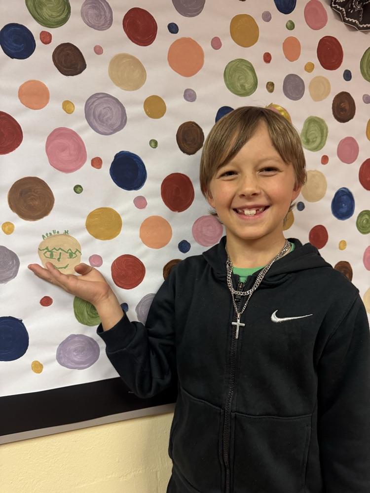 A smiling student proudly points to his artwork displayed on a colorful school bulletin board. His creative portrait is showcased among painted circles, highlighting student creativity and artistic expression.