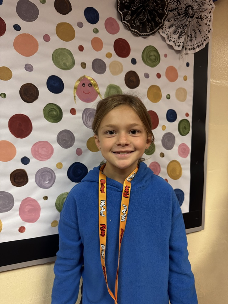 A smiling student proudly stands beside her artwork displayed on a colorful school bulletin board. Her creative portrait is featured among painted circles, celebrating her artistic expression and contribution to the display.