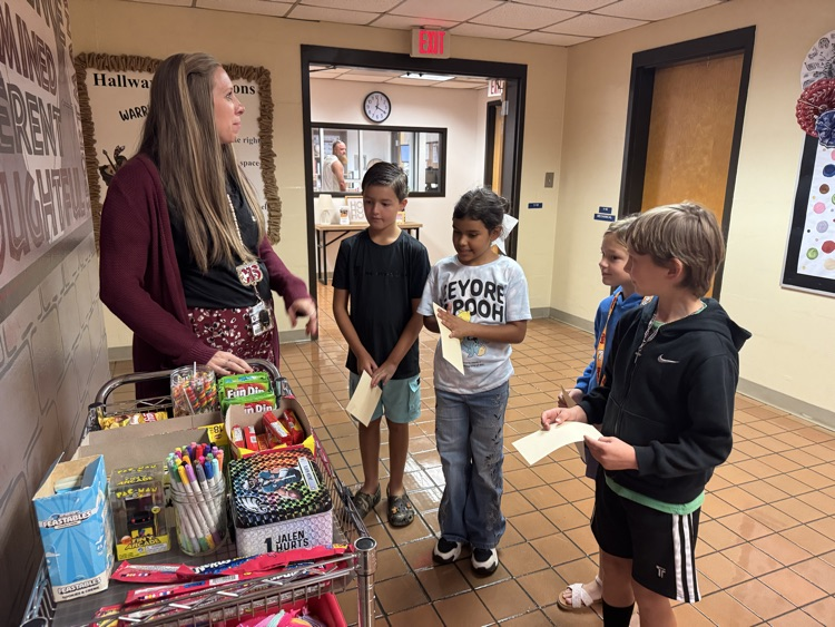 Ms.Simmons explains the prize choices to four students gathered around a cart filled with treats, toys, and colorful supplies. The students prepare to select rewards in celebration of their positive behavior and accomplishments.