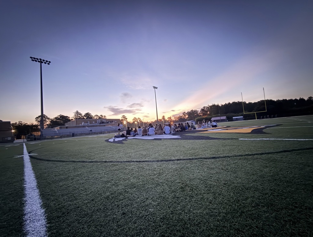 Three photos show a large group of West Nassau High School students gathered on the football field during an evening senior sunrise event. In the first photo, students pose together near the end zone as the sky begins to brighten. In the second and third photos, students sit together on blankets at midfield facing the sunrise, with the stadium, goalposts, and school buildings visible in the background