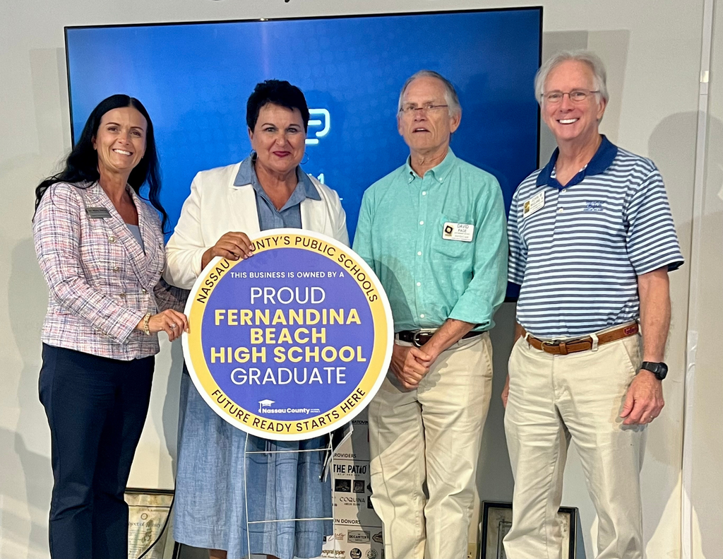 Four Fernandina Beach High School alumni stand together holding a Nassau County School District Alumni Business Recognition sign that reads, "Proud Fernandina Beach High School Graduate." Pictured are Executive Director of Curriculum & Instruction Dr. Misty Mathis, Superintendent Dr. Kathy Burns, Dr. David Page, and Dr. John McClane. The recognition celebrates Dr. Page's lifelong service to the Nassau County community as a family physician and Fernandina Beach High School graduate.