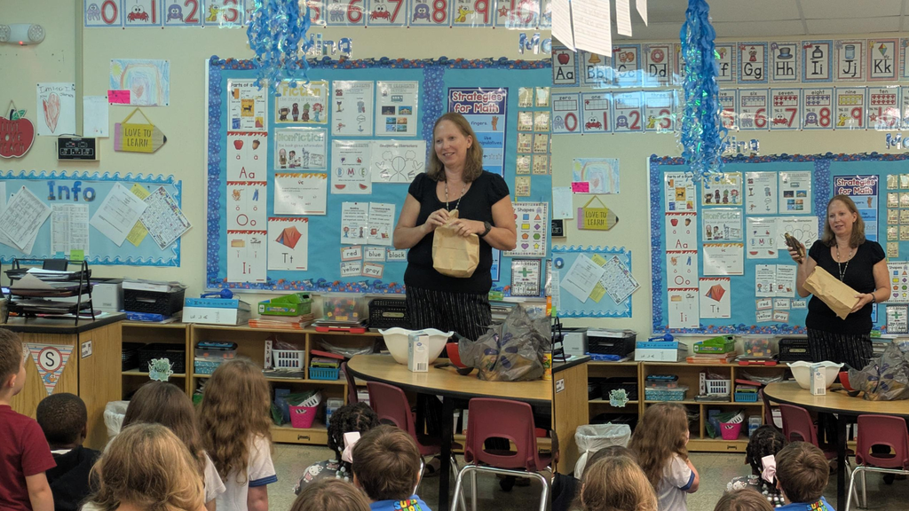 In a classroom, a teacher stands in front of a blue science-themed bulletin board with posters and charts while students sit at desks watching her presentation. Second picture: Classroom scene with a teacher standing at the front holding papers while students sit on the floor; a blue stream of glitter or confetti rises from a display board behind them.