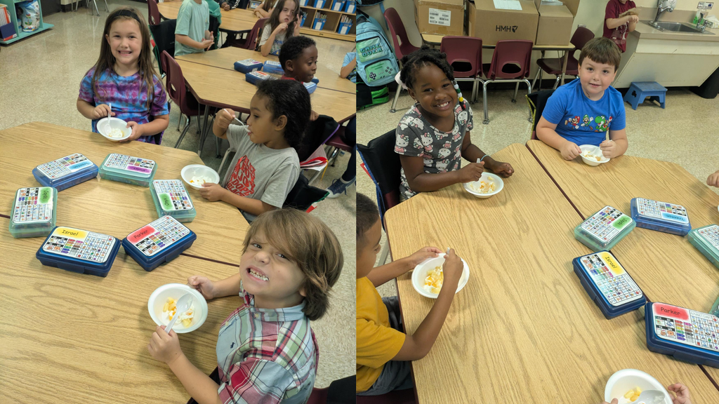 Children sit at desks in a classroom, smiling and eating from small white bowls while colorful learning kits with grid cards and bins are spread on the tables. Second picture: Two children sit at a classroom table eating from small plates, with colorful card games and a few blue containers on the tabletop and stacked boxes and chairs in the background.
