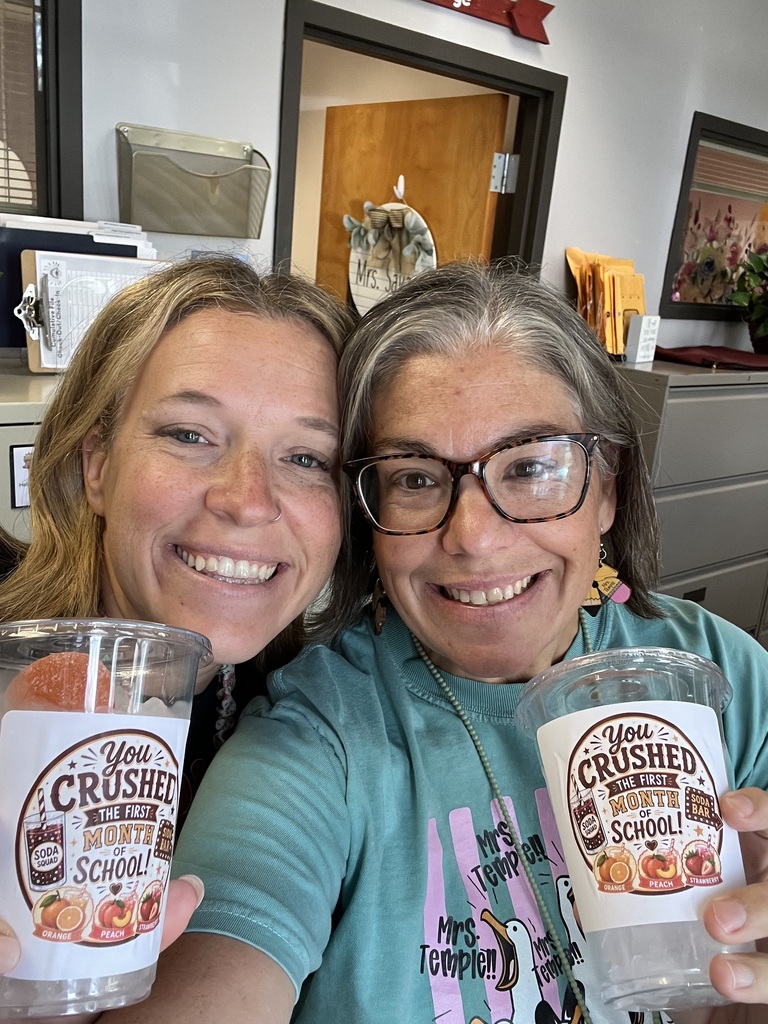 Two smiling school staff members pose closely together while holding clear cups featuring “You Crushed the First Month of School” labels. The labels include illustrations representing orange, peach, and strawberry flavors.