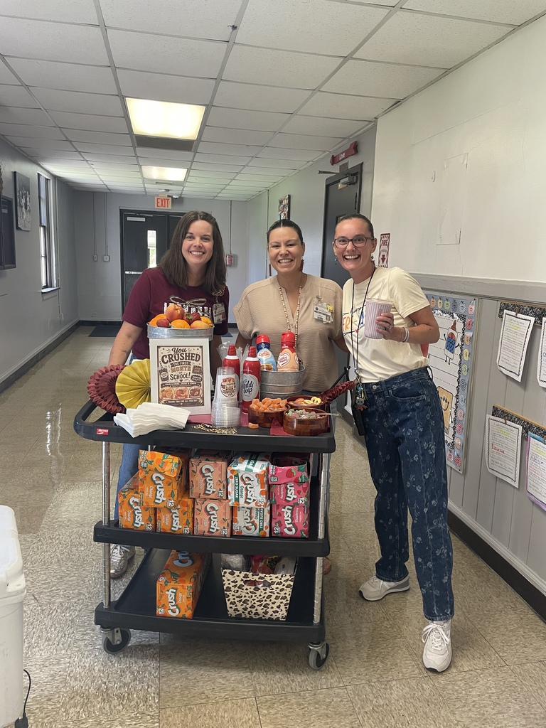 Three smiling school staff members stand beside a rolling cart decorated for a “You Crushed the First Month of School” celebration. The cart is stocked with orange, peach, and strawberry soda, whipped cream, fruit, toppings, cups, and napkins. A sign on the cart promotes the celebration.