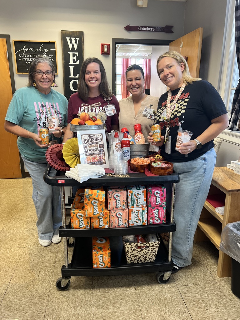 Four smiling school staff members pose behind a decorated beverage cart while holding drinks. The cart is filled with orange, peach, and strawberry Crush sodas, whipped cream, fruit, toppings, and supplies for the staff celebration.