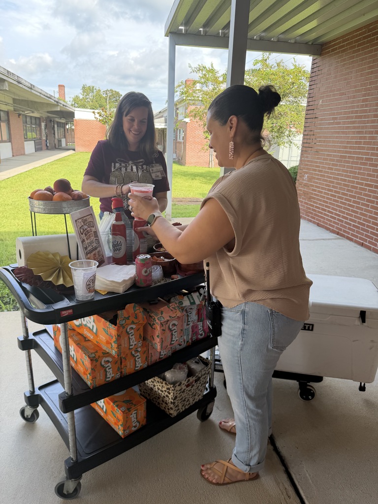 Two school staff members prepare drinks at a decorated beverage cart outdoors on a school campus. The cart is stocked with Crush sodas, whipped cream, fruit, toppings, cups, and napkins, with a “You Crushed the First Month of School” sign displayed.