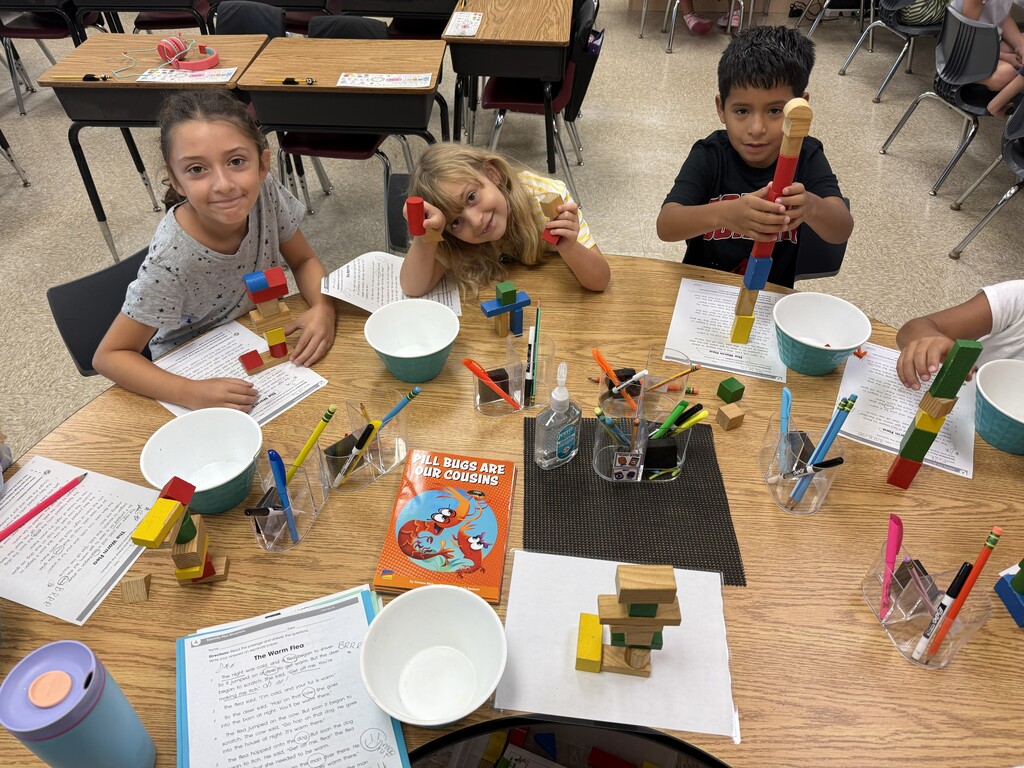 Three children sit around a wooden table with bowls, colored building blocks, and craft materials, holding small red and blue pieces while working on a classroom activity.