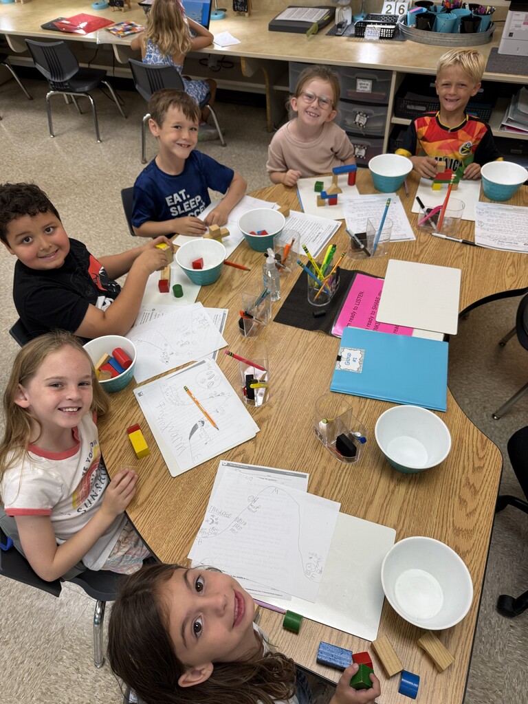 A classroom group sits around a wooden table working on plot structure using building blocks to represent the plot. Passages students are reading in front of them.