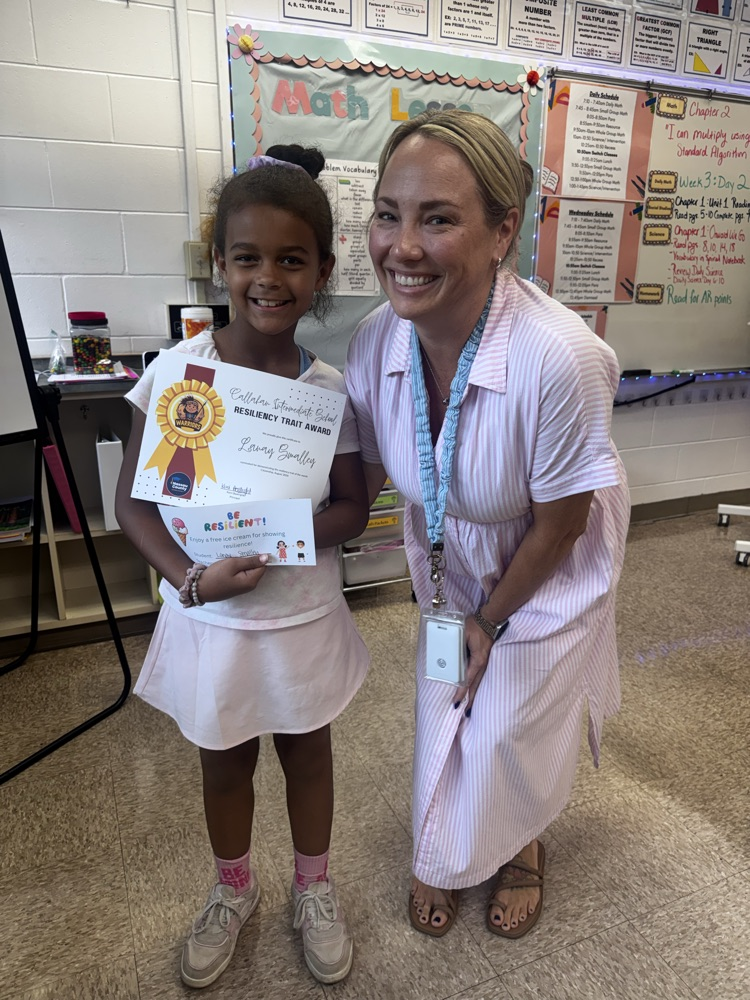 A smiling elementary student stands beside her teacher in a colorful math classroom while proudly holding a Callahan Intermediate School “Resiliency Trait Award” certificate and a “Be Resilient!” recognition card. Both are smiling at the camera.