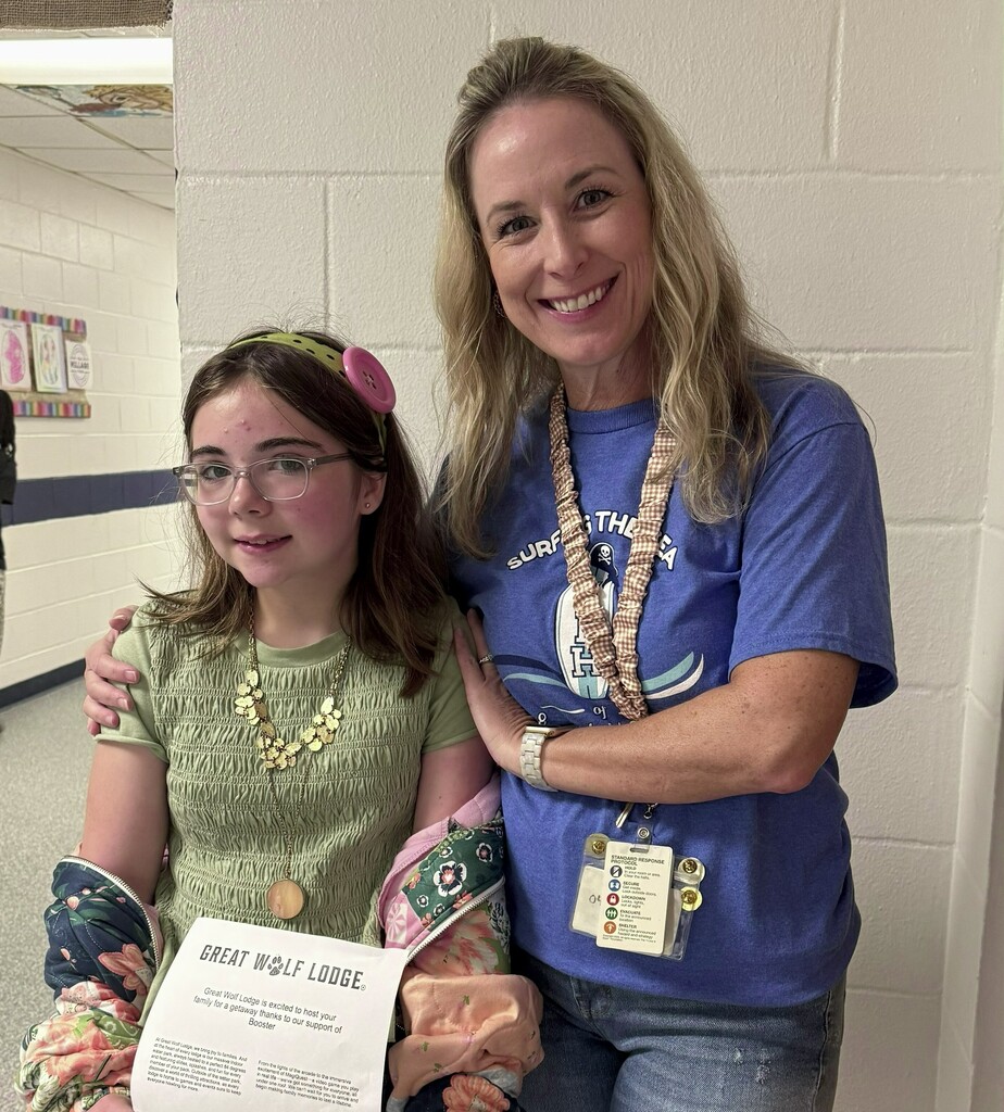 A girls holding a Great Wolf Lodge certificate smiling.  Her teacher is smiling as well and standing beside her with her arms on her shoulders. 