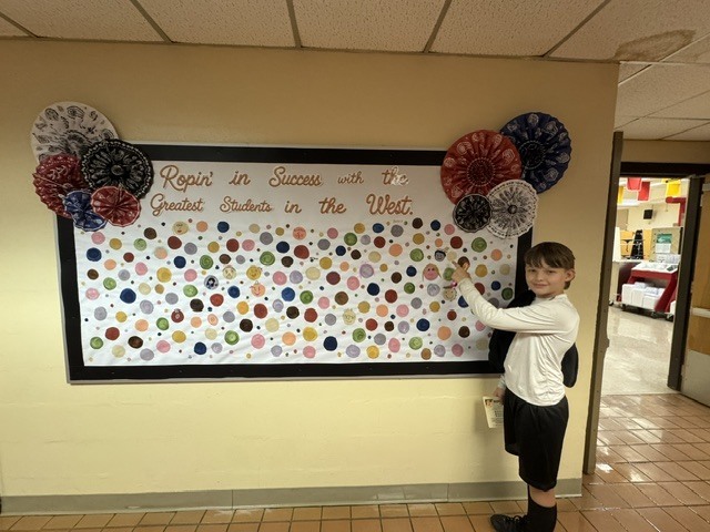 A student proudly points to their artwork dot displayed at school. The student smiles while showcasing their creativity and hard work.
