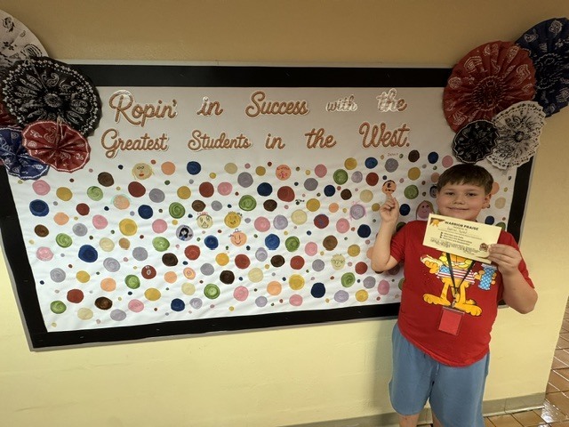 A student proudly points to their artwork dot displayed at school. The student smiles while showcasing their creativity and hard work.