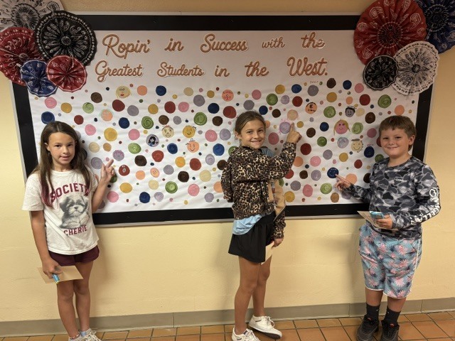 Students proudly point to their dots representing our school-wide expectations: Be Respectful, Be Responsible, and Be Safe. The students smile as they show their commitment to making positive choices at school.