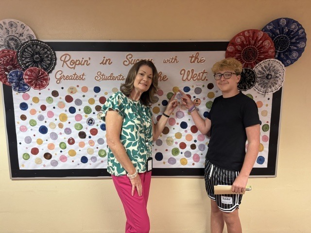 A student proudly points to their artwork dot displayed at school. The student smiles while showcasing their creativity and hard work.