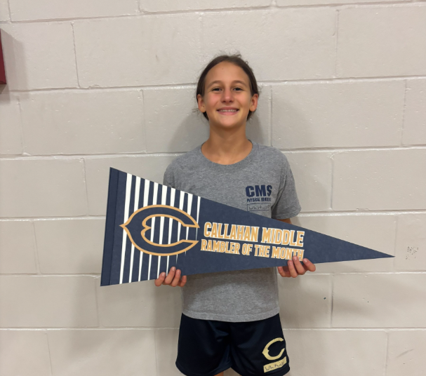 Student standing against a white cinderblock wall holding a blue and gold pennant that reads “Callahan Middle Rambler of the Month.” The student is wearing a gray CMS T-shirt and athletic shorts.