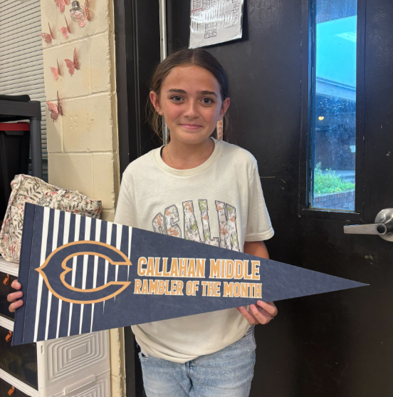 Student standing in a classroom holding a blue and gold pennant that reads “Callahan Middle Rambler of the Month.” A classroom door, storage containers, and wall decorations are visible in the background.