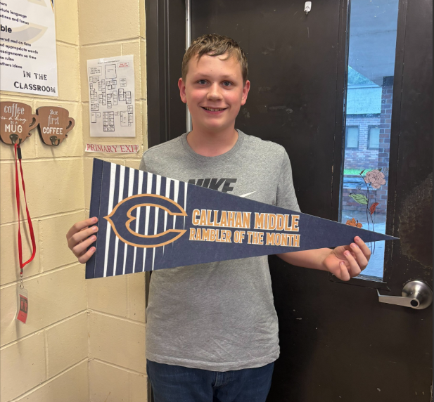 Student standing in a classroom holding a blue and gold pennant that reads “Callahan Middle Rambler of the Month.” A classroom door, wall posters, and school signage are visible in the background.