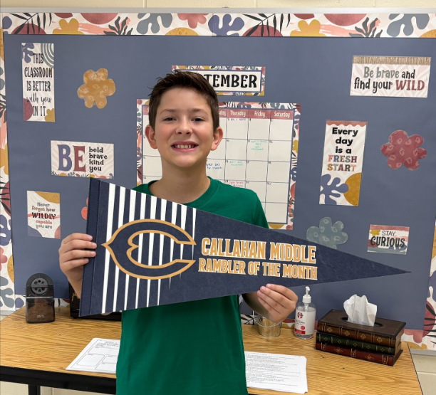 Student standing in a classroom holding a blue and gold pennant that reads “Callahan Middle Rambler of the Month.” A September calendar and classroom motivational posters are displayed on the bulletin board behind the student.
