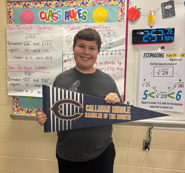 Student standing in front of a classroom math bulletin board holding a blue and gold pennant that reads “Callahan Middle Rambler of the Month.” Math rules, square root examples, and a digital classroom clock are displayed behind the student.