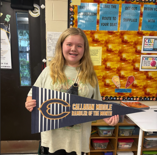 Student standing in a classroom holding a blue and gold pennant that reads “Callahan Middle Rambler of the Month.” Classroom rules posters, storage bins, and a bulletin board are visible in the background.