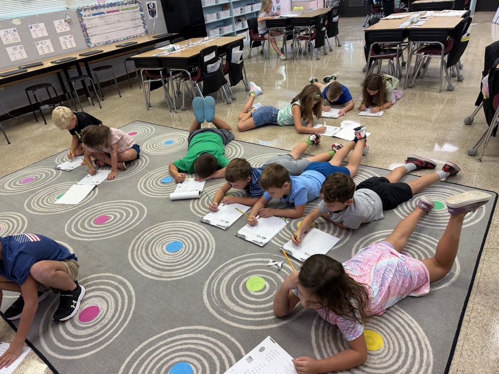 multiple students laying on a gray carpet with clipboards working on school work