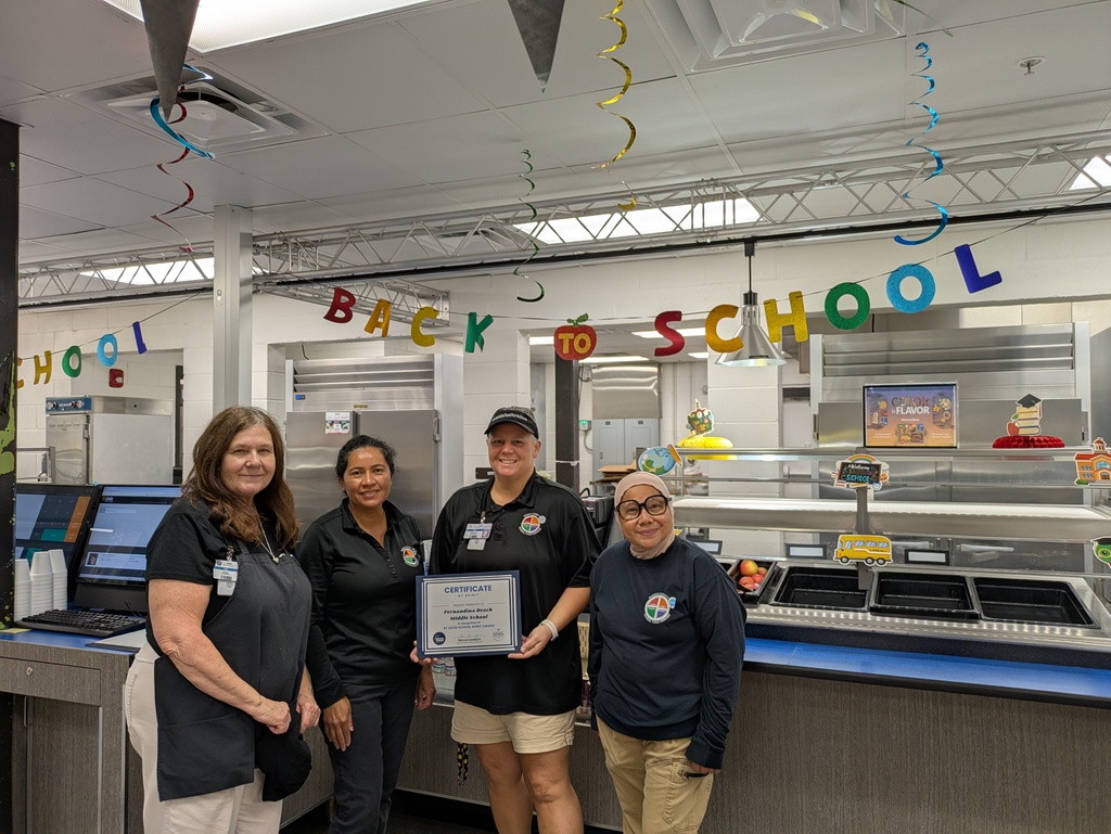 Picture of four cafeteria workers posing with their award in front of the cafeteria line and a "Back to School" banner.