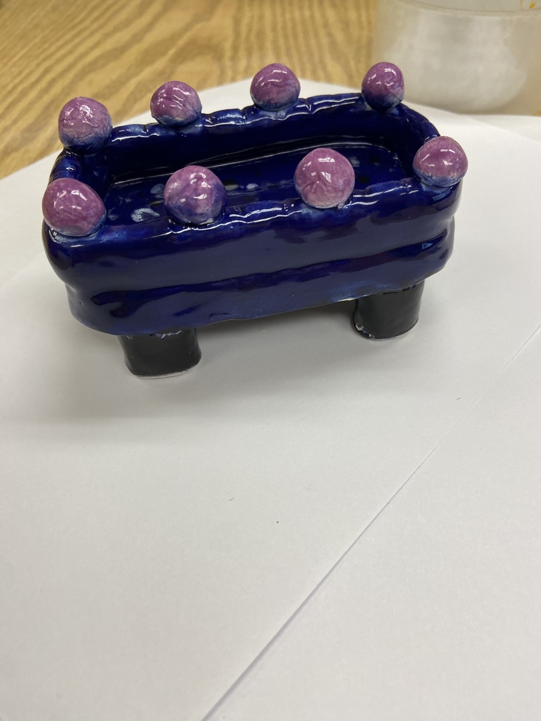 Picture of sample student ceramic artwork: Blue glazed ceramic container with pink-purple spherical accents around the rim and four short legs.