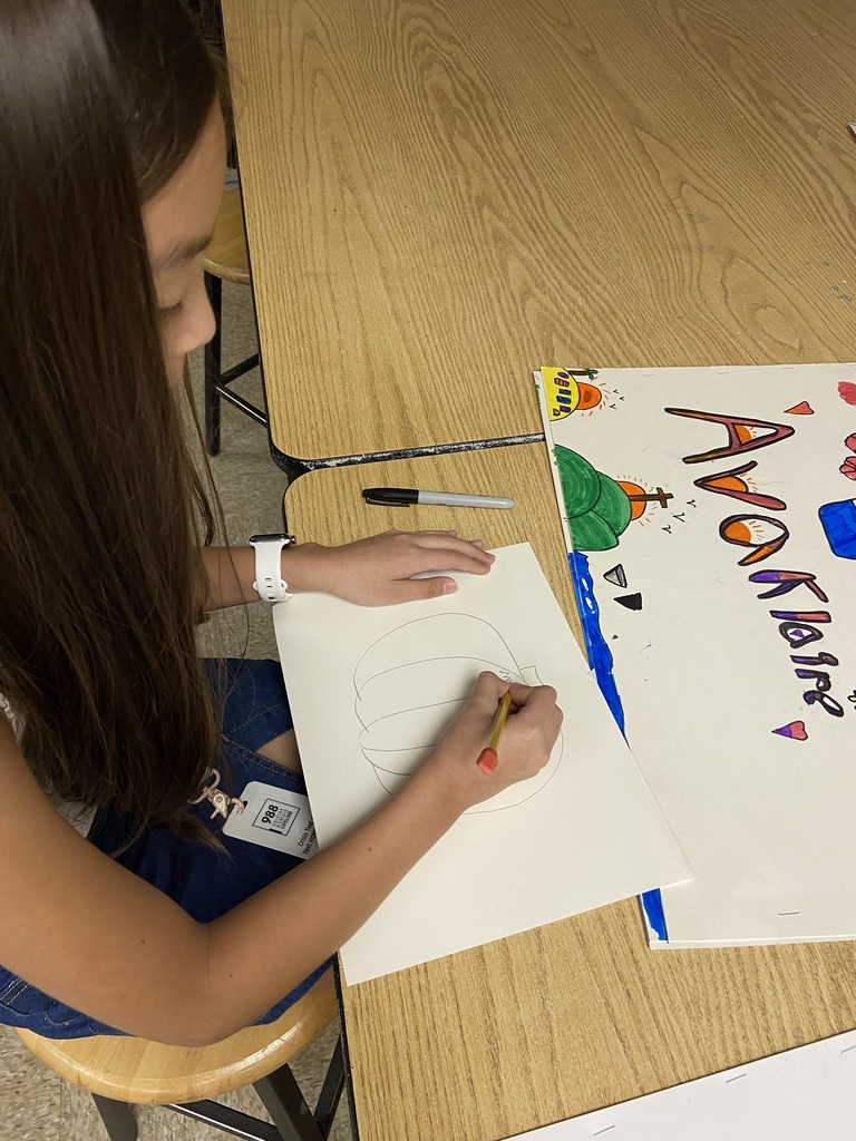 Student sketching a pumpkin drawing with a pencil at a classroom table.