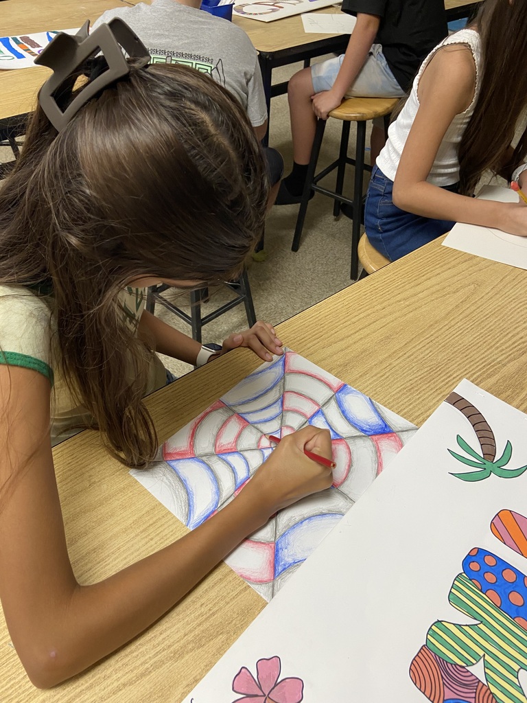 Student creating a geometric optical illusion drawing with red, blue, and gray shading at a classroom table.